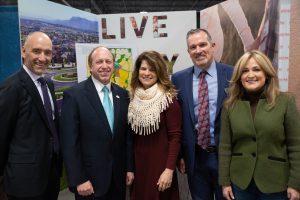 5 people including John Suthers standing in front of miscellaneous posters